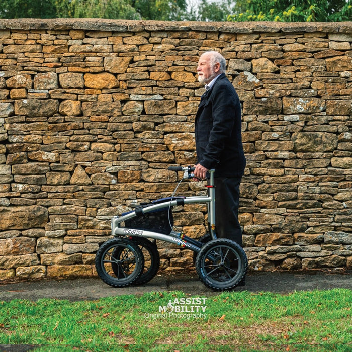 Side view of the Trionic Veloped Sport rollator and its large all-terrain wheels, with an older man against a stone wall