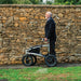 Side view of the Trionic Veloped Sport rollator and its large all-terrain wheels, with an older man against a stone wall