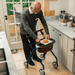 An older man in a kitchen uses his Trust Care Let's Dream indoor rollator with a tray . The rollator has a dark blue frame, brown leather-like accents, and black wheels.