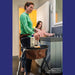 A woman in a green sweater uses a Trust Care Let's Dream indoor rollator in a bathroom while a young girl stands at the sink. The rollator has a brown front storage bag and tray holding toiletries. 