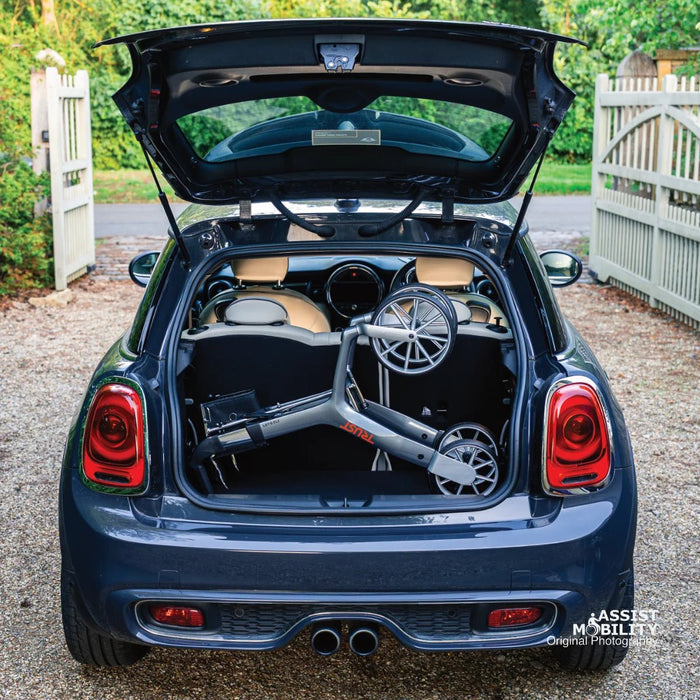 The compact folded Trust Care Let's Fly rollator in the boot of a dark blue Mini Cooper on a gravel driveway