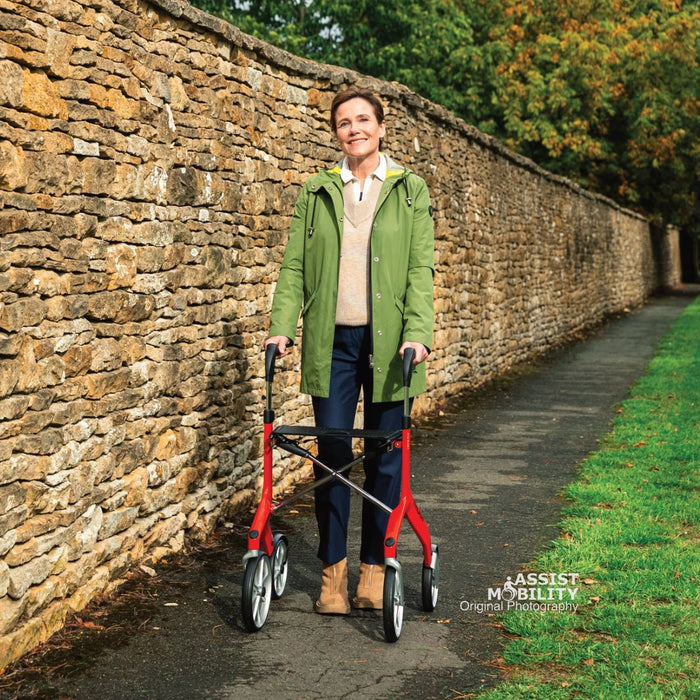 Smiling woman standing with the red Trust Care Let's Fly rollator on a path between a stone wall and autumn trees