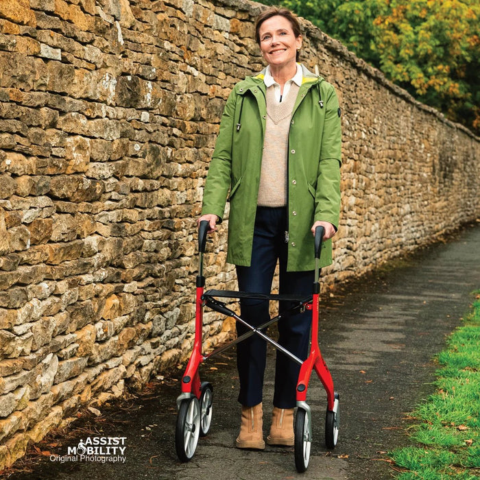 Smiling woman standing tall with the red Trust Care Let's Fly rollator on a path beside a Cotswold stone wall