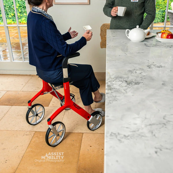 A person in a navy sweater sits on a red Trust Care Let's Fly rollator with a seat, positioned in a bright kitchen next to a marble counter.
