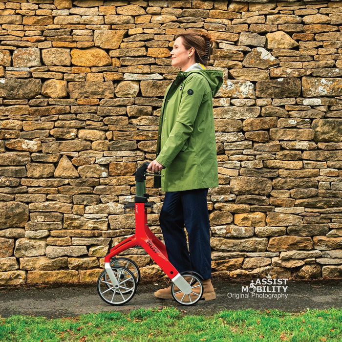 Side-on view of the red Trust Care Let's Fly rollator with a woman in a green coat against a dry stone wall