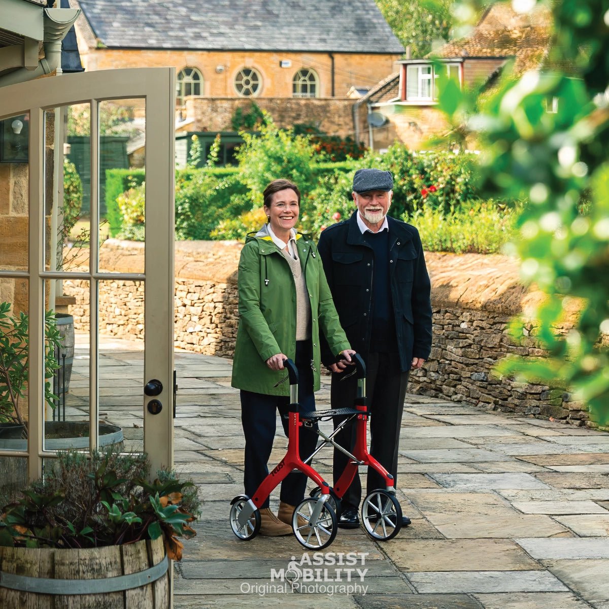 An older man and woman standing outdoors on a stone path in front of a stone building in the Cotswold. The woman is using a red Trust Care Let's Fly rollator, and the man is standing beside her.