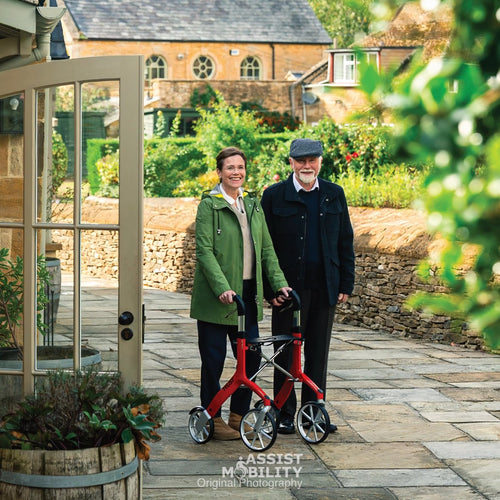 An older man and woman standing outdoors on a stone path in front of a stone building in the Cotswold. The woman is using a red Trust Care Let's Fly rollator, and the man is standing beside her.