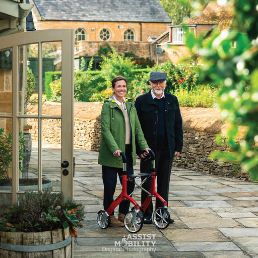 An older man and woman standing outdoors on a stone path in front of a stone building in the Cotswold. The woman is using a red Trust Care Let's Fly rollator, and the man is standing beside her.