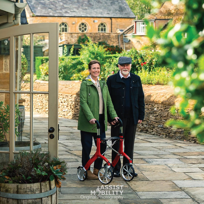 An older man and woman standing outdoors on a stone path in front of a stone building in the Cotswold. The woman is using a red Trust Care Let's Fly rollator, and the man is standing beside her.