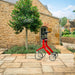 A red Trust Care Let's Fly rollator, positioned on a stone patio in front of a stone cottage with a green tree in the Cotswolds.