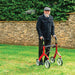 Older man standing with a red Trust Care Let's Fly rollator and its built-in seat on a lawn beside a Cotswold stone wall