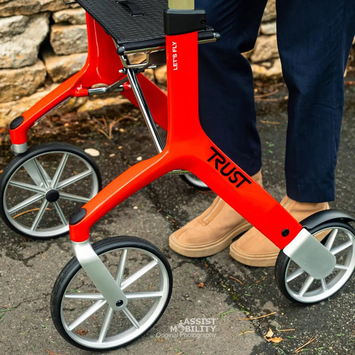 Close-up of a bright red Trust Care Let's Fly rollator with silver wheels, shown outdoors next to a person wearing navy trousers and tan boots.