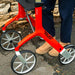 Close-up of a bright red Trust Care Let's Fly rollator with silver wheels, shown outdoors next to a person wearing navy trousers and tan boots.