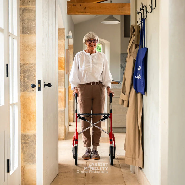 An older woman stands in a hallway using a red Trust Care Let's Fly rollator. She is wearing a white blouse, brown trousers, and glasses.