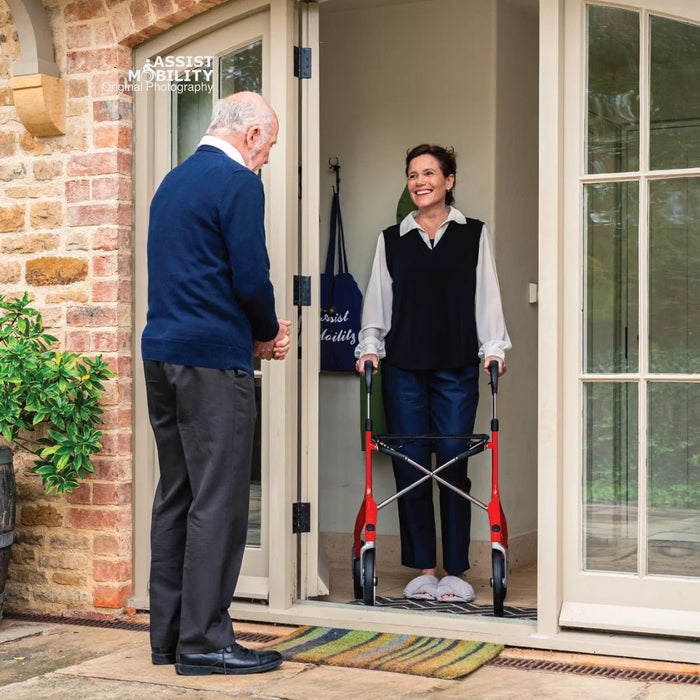 Smiling woman at her front door with a red Trust Care Let's Fly rollator, welcoming an older male visitor