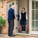 Smiling woman at her front door with a red Trust Care Let's Fly rollator, welcoming an older male visitor