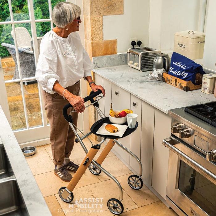 An older woman in a white blouse and brown trousers stands in a bright kitchen using a wooden and Trust Care Lets Go Indoor rollator with a tray and cup on it. The scene includes a toaster, kettle, bread box, and stove.