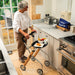 An older woman in a white blouse and brown trousers stands in a bright kitchen using a wooden and Trust Care Lets Go Indoor rollator with a tray and cup on it. The scene includes a toaster, kettle, bread box, and stove.