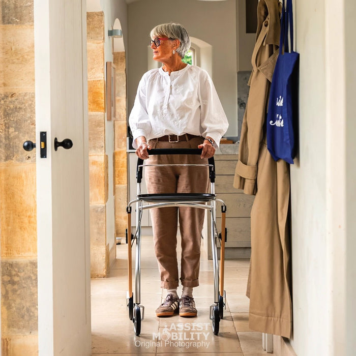 An older woman with grey hair and glasses pushes a Trust Care Lets Go Indoor rollator down a hallway. She is wearing a white button-down shirt and brown trousers.