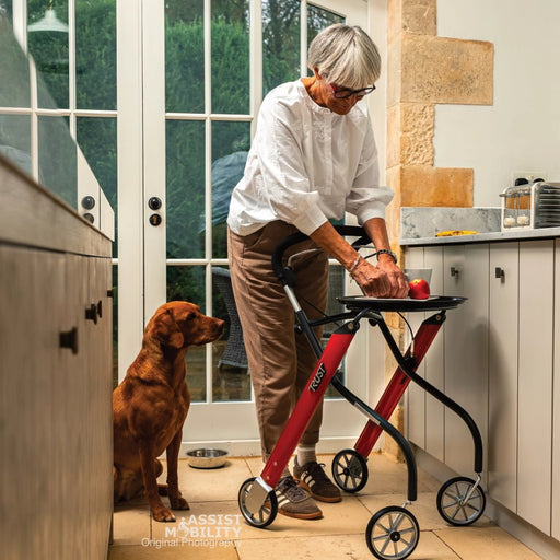 An older woman using a red and black Trust Care Lets Go Indoor rollator  in a kitchen, with a brown dog sitting nearby.