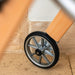 A close-up of the rear wheel on a Trust Care Let's Go Indoor rollator, positioned against a beige stone and white wall background.