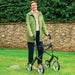 Woman smiling with her British Racing Green Trust Care Let's Go Out rollator, showing its seat and large wheels on a lawn