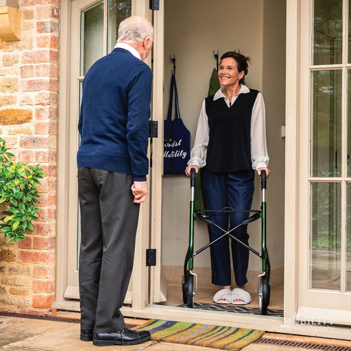 An elderly woman stands at the doorway of a home using a green Trust Care Let's Go Out rollator, while an elderly man in a navy sweater and grey trousers stands outside looking at her. A navy tote bag with white text hangs on the door.