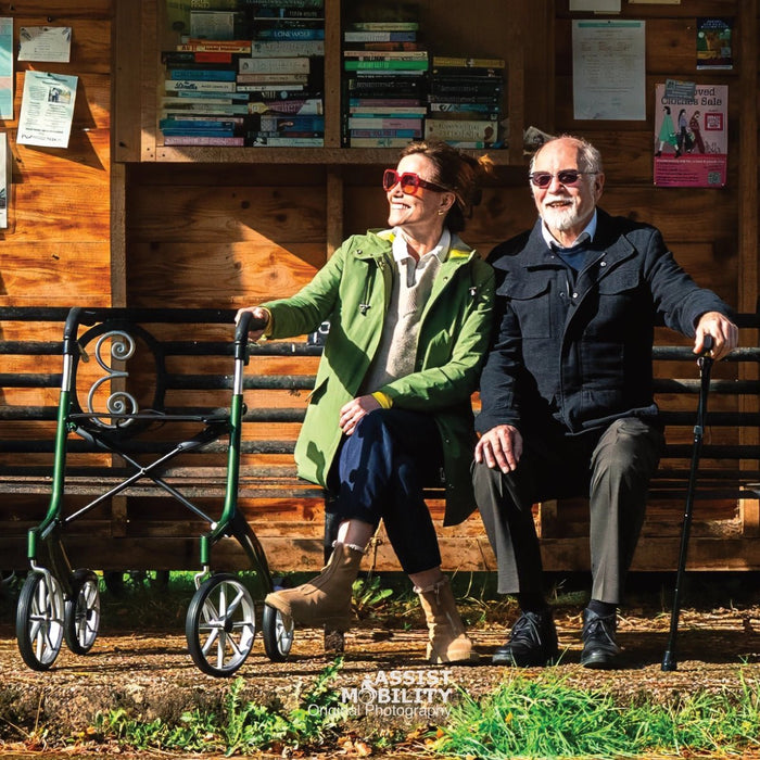 An elderly couple sits on a wooden bench at the bus top, with the woman using a green Trust Care rollator and the man holding a cane. The woman wears a green coat and sunglasses, and the man wears a dark jacket and sunglasses.
