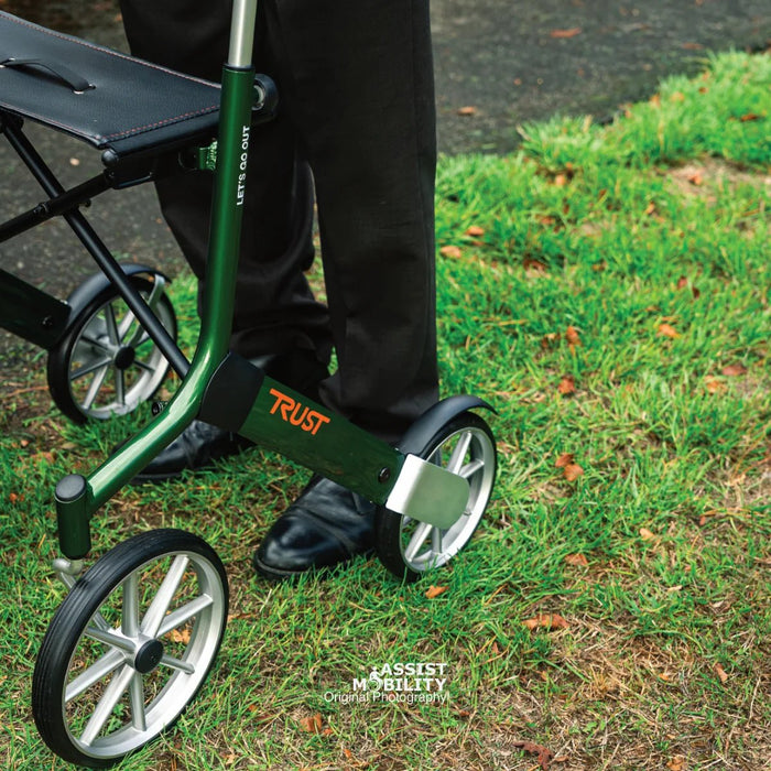Close-up of the British Racing Green Trust Care Let's Go Out rollator frame, showing the Trust logo, seat and spoked wheels on grass