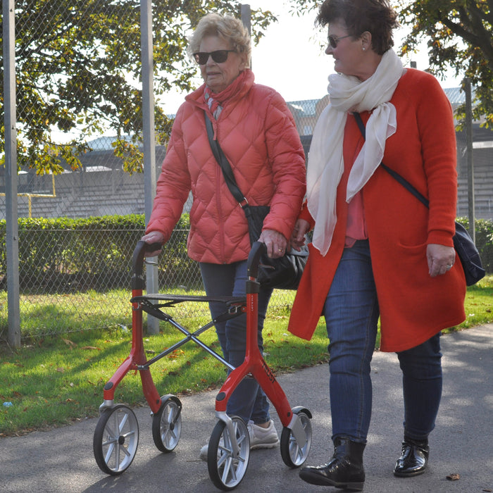 Two ladies walking side by side with a Red Lets Fly rollator with colour cordinated coats.